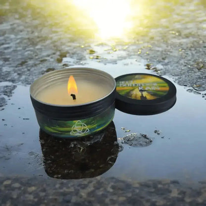 Lit Irish Summer Rain tin candle standing in a shallow puddle, lid beside it